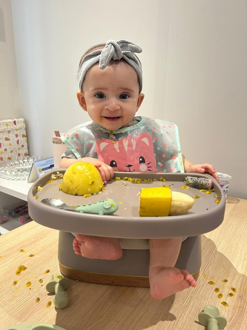 Bebê sorridente na cadeira de alimentação explorando frutas como maracujá e banana com as mãos durante processo de introdução alimentar.