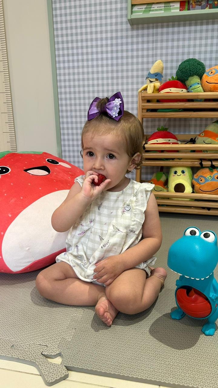 Criança pequena sentada no tapete do consultório comendo um morango, cercada por pelúcias de frutas e brinquedos educativos sobre alimentação saudável.