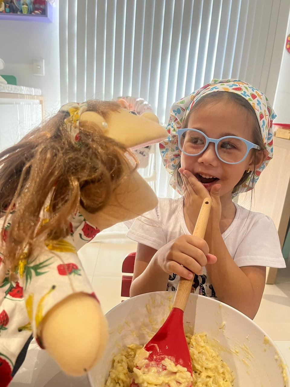 Criança com óculos e touca de chef rindo e interagindo com um fantoche de mão divertido durante uma oficina culinária infantil focada em educação alimentar lúdica.